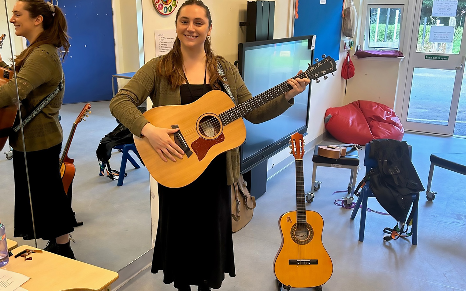 Musician Liv Dawn stands in a brightly lit room, with a guitar in her arms, and one in a stand on the floor next to her. Around her are tall mirrors, a large TV, bean bags, instruments and desks.