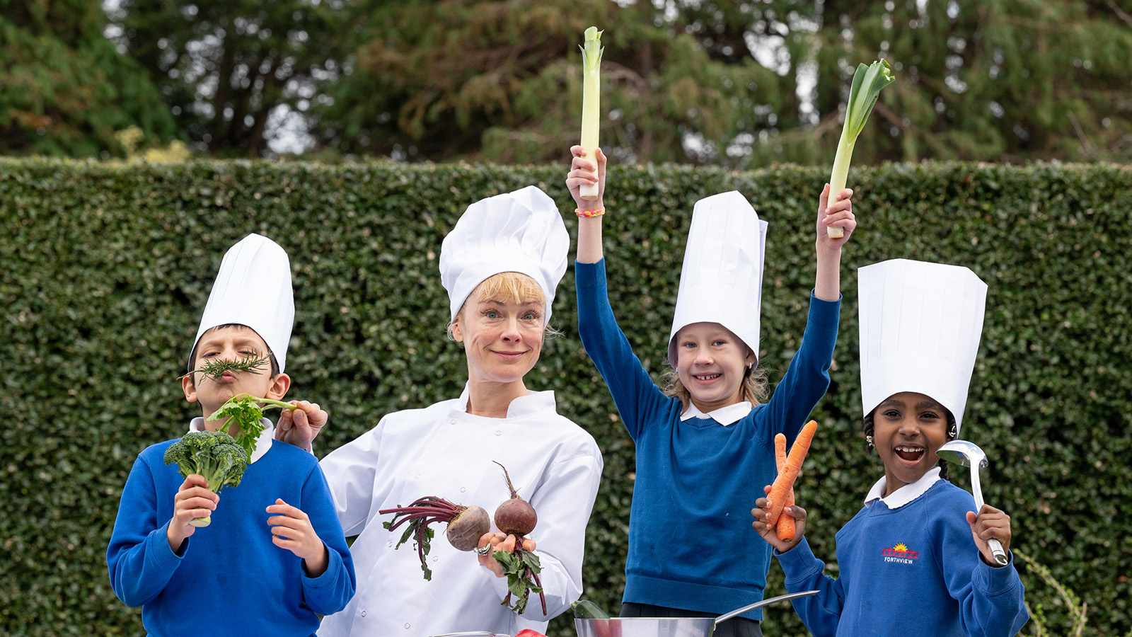 A woman with blonde hair wearing a traditional white chef's outfit and hat and three young children in bright blue jumpers hold up various vegetables