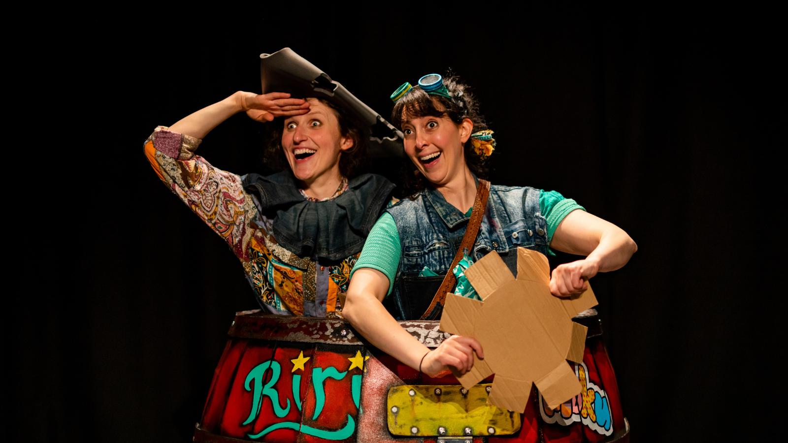 Two performers stand on stage. They look as though they are pirates, with one holding her hand up to her face as though she is looking towards land