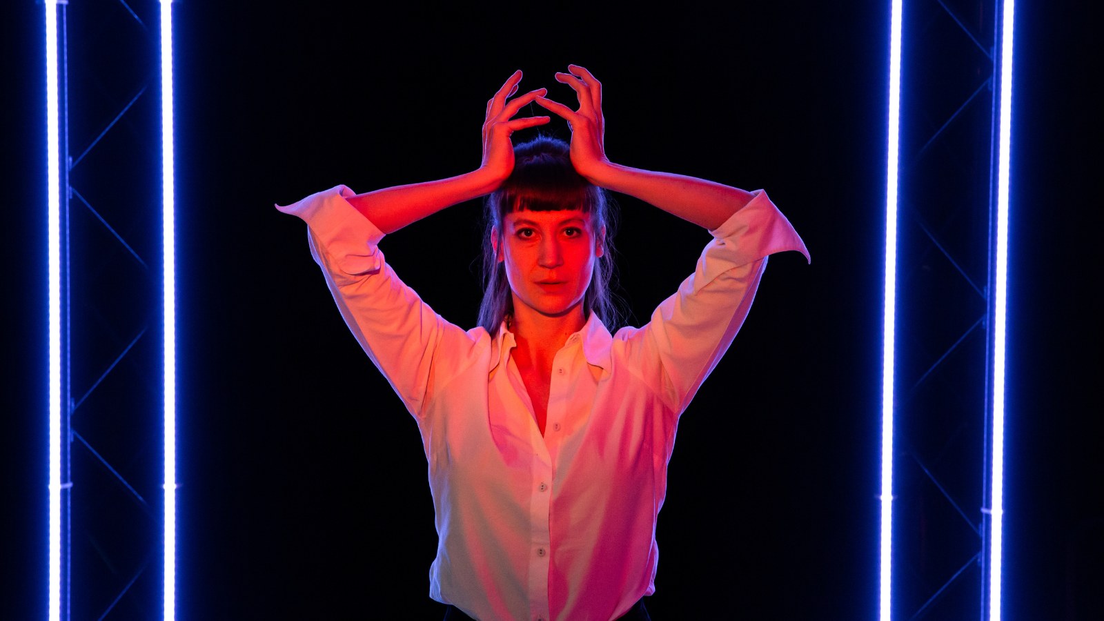 A woman stands in a white shirt flooded with red light. She makes a crown shape above her head with her hands.