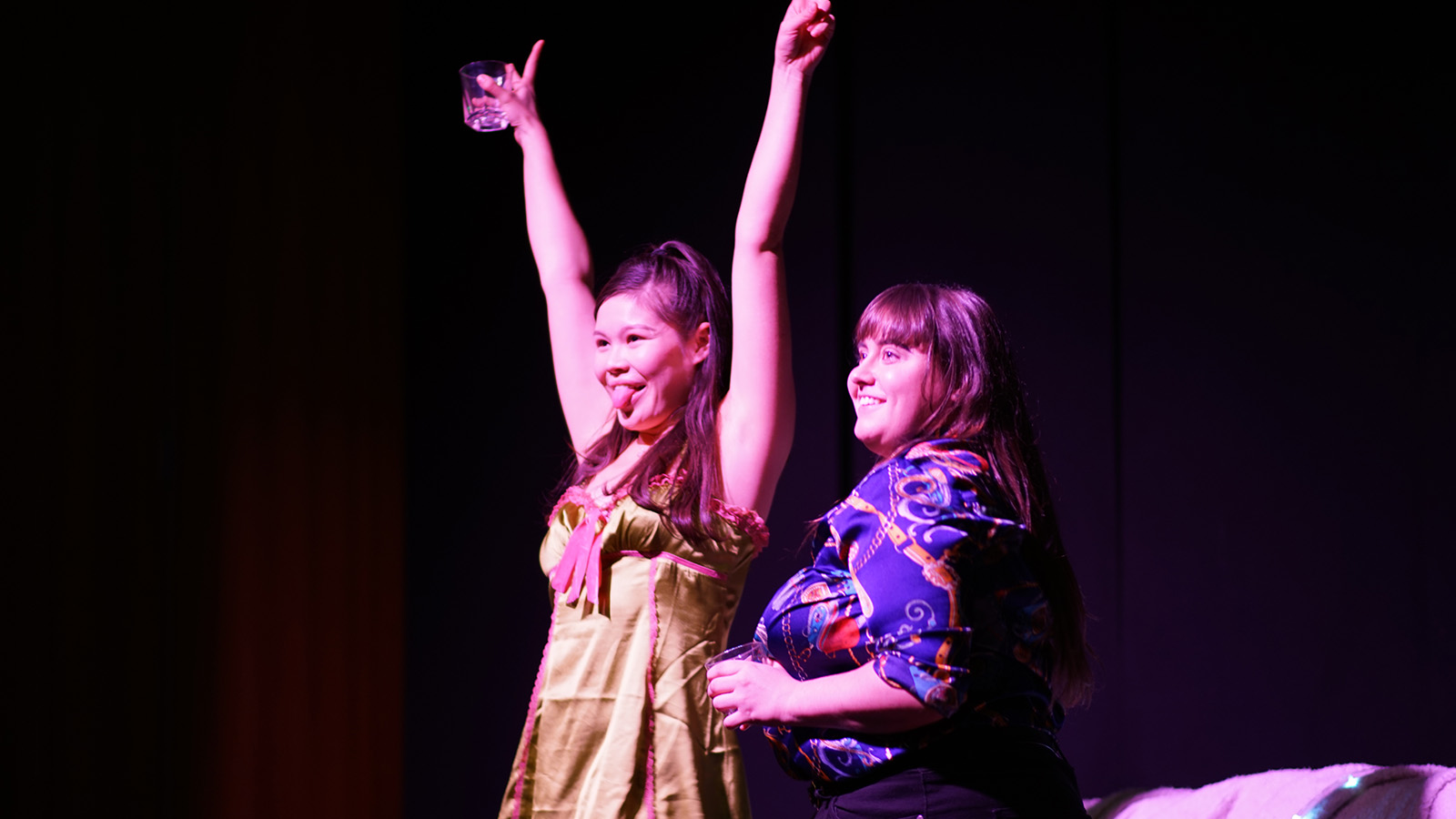 Two women stand on stage, one with her arms in the air and her tongue out