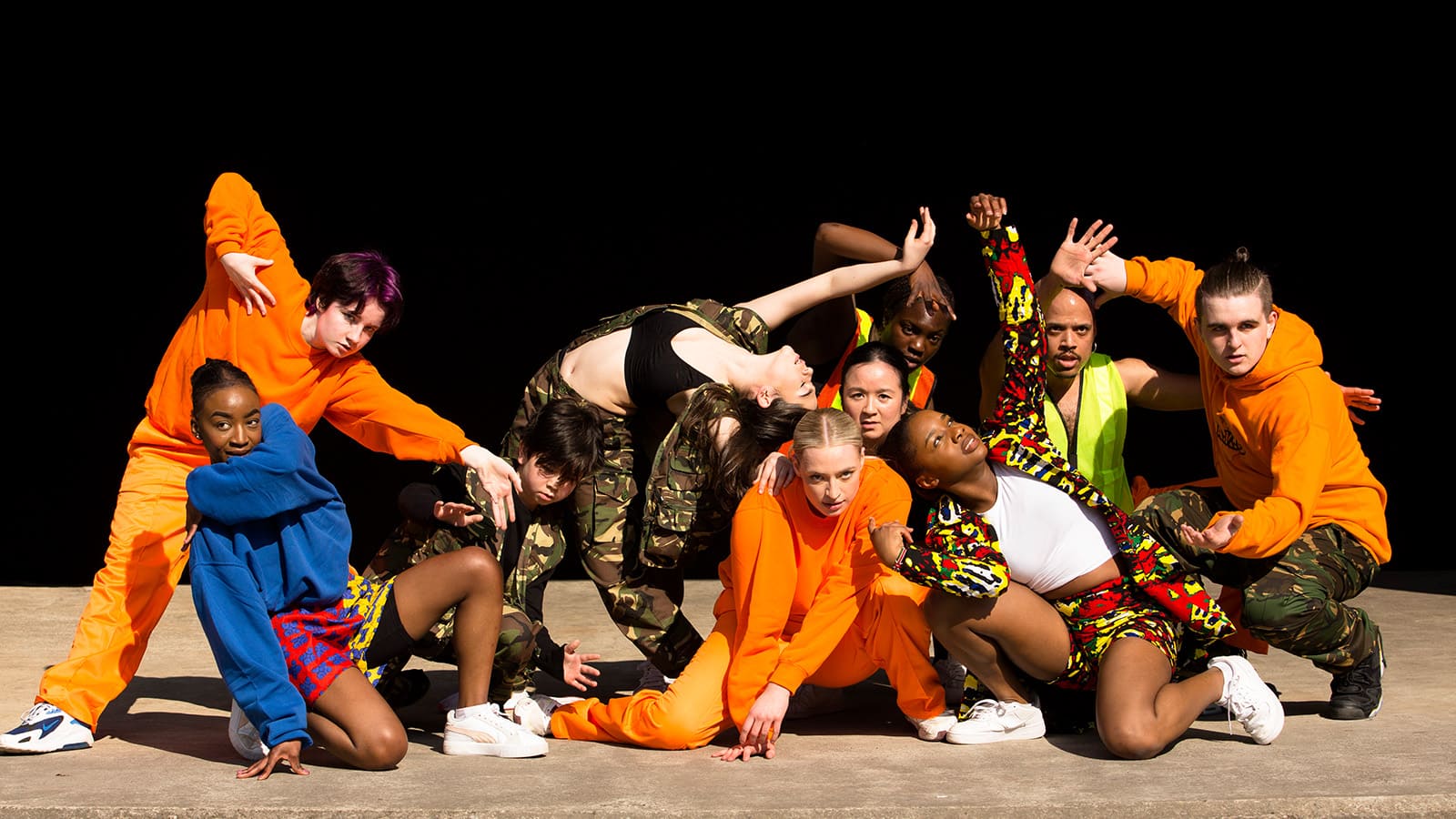 A group of dancers pose on a stage, all dressed in camouflage, or bright orange, green and red