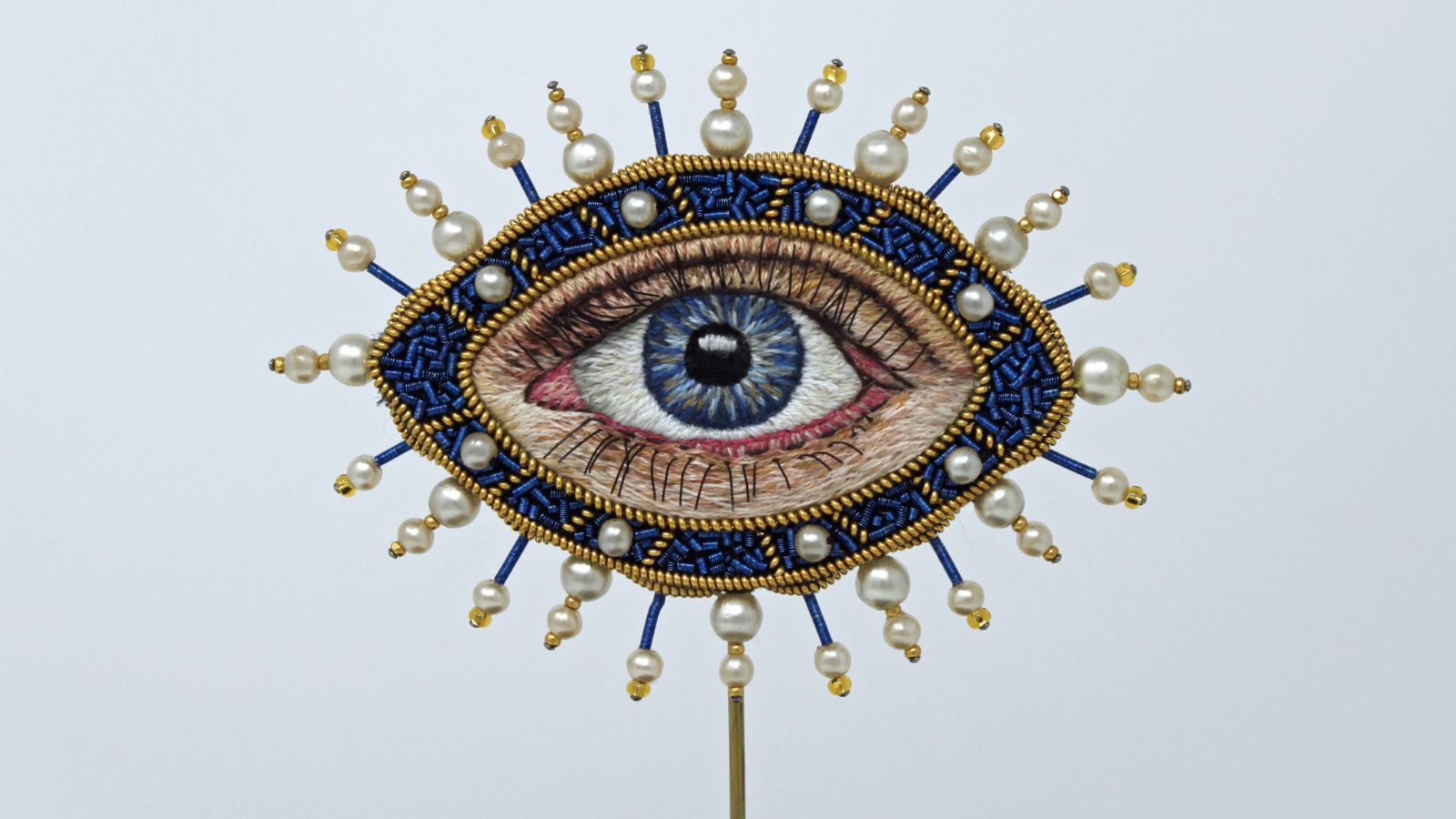 A highly detailed embroidered blue eye with gold spokes sticking out all around it by artist Tzipporah Johnston