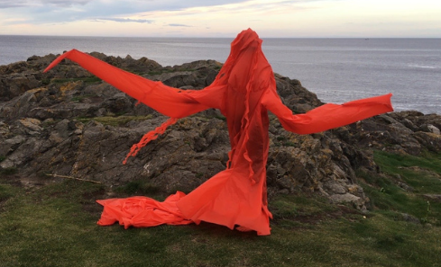 A figure stands on a beach in a bright red angular costume made of recycled plastics