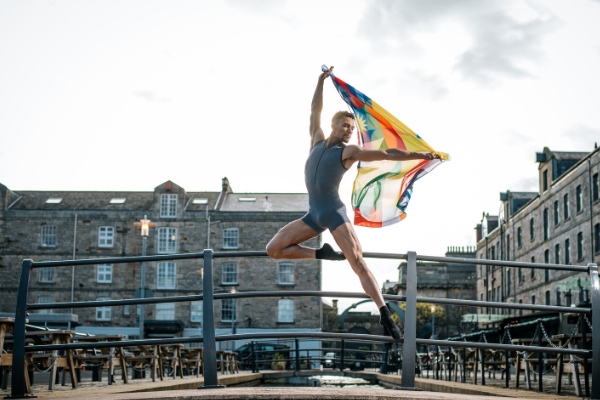 A dancer holding a rainbow flag leaps into the air - he looks very physically strong and is wearing a unitard
