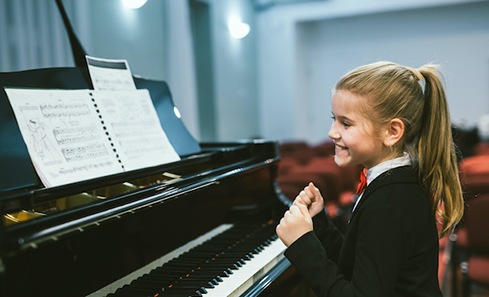 A girl in a school uniform sits in front of a piano looking excited