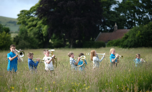 Young children playing brass instruments stand in a field of flowers on a bright and sunny day