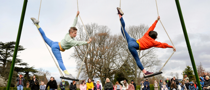Two performers from All or Nothing balance on swings high in the air outside as an audience watches on