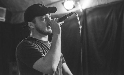 Local hip hop artist Jackill stands holding a microphone in a black and white image - he wears a black cap and looks away from the camera