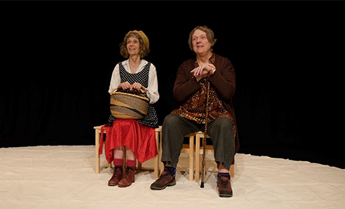Image shows two people sitting on wooden chairs on a stage with a black backdrop. They are both looking out at the audience. One has a wicker basket on her knee and the other is holding a wooden cane. 