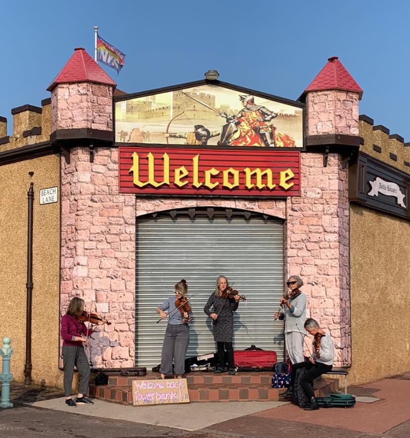 A group of fiddle players stand outside on a sunny morning under a sign that says welcome
