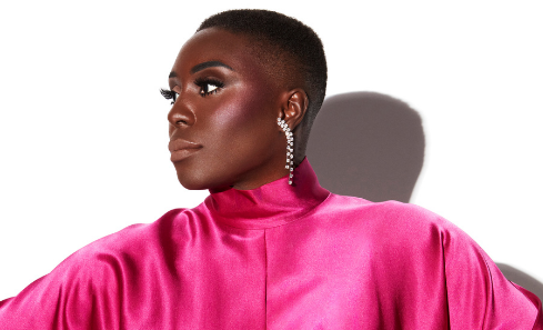 Laura Mvula stands in a vibrant pink dress against a high contrast white background, looking powerful as she looks to the left of the camera