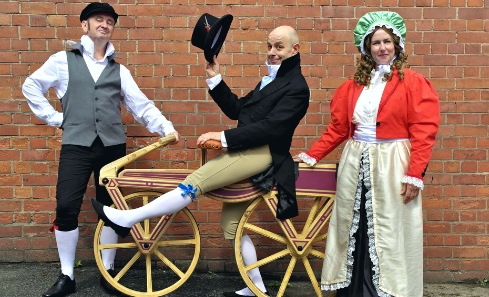 Three people in period dress stand in front of a brick wall. One of them straddles a bicycle with his hat jauntily thrown in the air. They look as though they are from the 1600s.