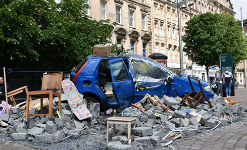 An installation of a destroyed car in the middle of a leafy street