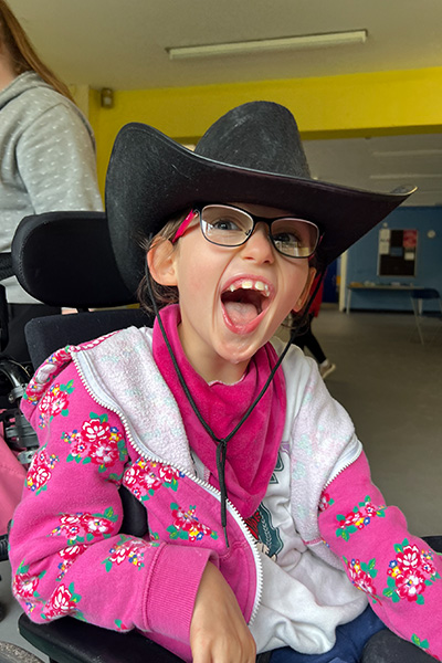 A young girl in a wheelchair wearing glasses and a bright pink kerchief on her neck and a black cowboy hat. She has a big smile on her face.