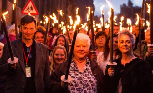 Three people stand in front of a torch lit procession crowd.