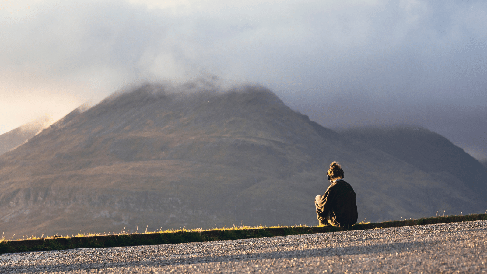 A person sits at the side of the road looking out over a hill that has clouds nestled at the top of it - they are wearing headphones