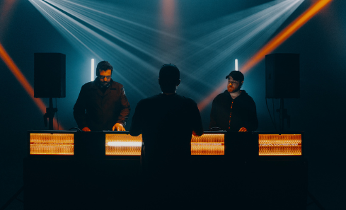 A group of people stand in front of turntables in a dark club with orange lights
