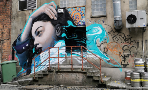 A colourful piece of street art in Glasgow by the artist Mark Worst. A woman brushes her hair back from her face, swirls of blue and white surround her as if her hair has transformed. The street art stands out against a grey and brown background of doors, walls, windows and vent.