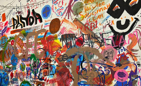 A white wall has been covered in a variety of wild, colourful and playful drawings, illustrations and markings. One is a face with the tongue sticking out, many are simple wipes, scrapes and lines, others are letters, faces and words. It’s a visual cacophony. 