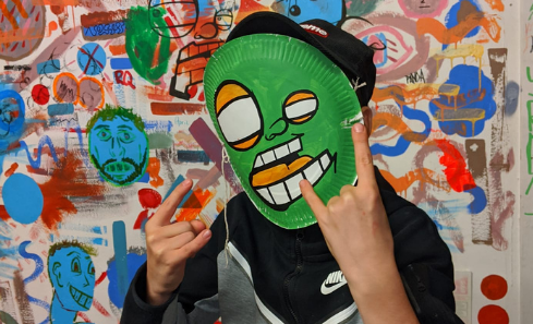 A young man wearing a green mask with a cartoon face on it makes the devil horns hand sign at the camera Behind him is a white wall covered in colourful and playful drawings, illustrations and markings. It’s a visual cacophony