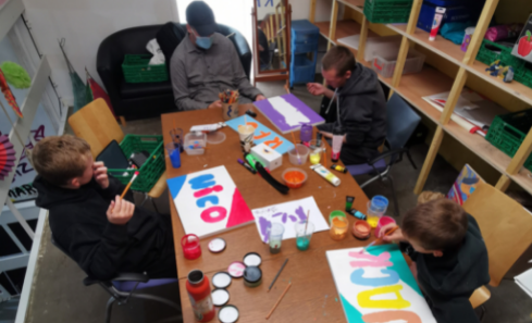 Panda and three young people sit around a table at a Colour Ways workshop at G20. The table is covered in drawings and art supplies. They’re working on their own pieces of art, holding paintbrushes, with pots at their sides filled with red, blue, yellow and orange paint.