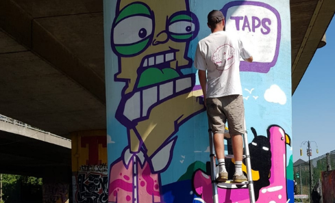 The artist Panda stands at the top of a ladder leaning against a concrete pillar. He’s painting a street art mural of a hot and sunny day. It shows a cartoon man standing against a clear blue sky. In the background of the mural there’s a character floating in an inflatable. Panda appears to be drawing the words ‘taps aff’.
