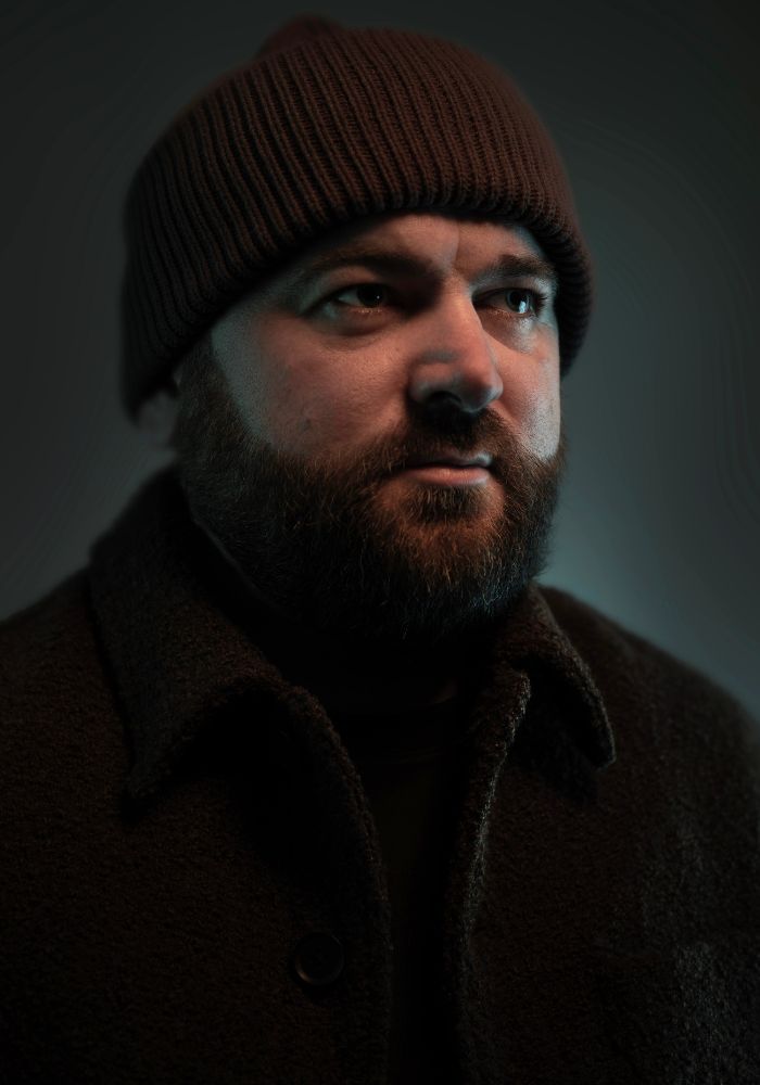 A person wearing a dark knit hat and a textured coat stands in low light, creating a mysterious atmosphere.