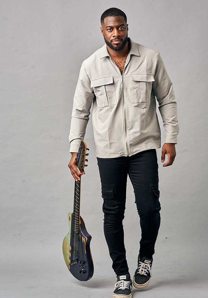 A person in a light gray zip-up jacket with two front pockets holding a Hybrid guitar, posed against a muted gray background.