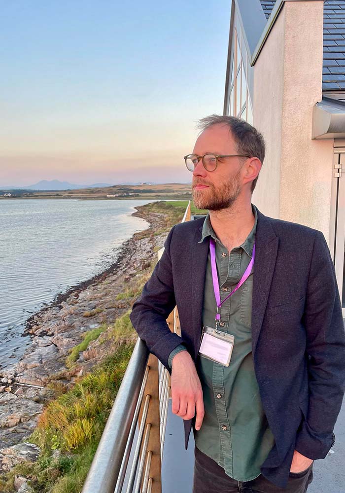 A person in a dark blazer and green shirt stands by a railing, overlooking a scenic shoreline and a calm body of water at sunset.