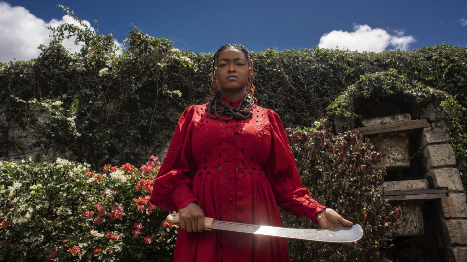 A woman stands proudly wearing a red dress and holding a large curved knife outside in front of a lush garden against a blue sky