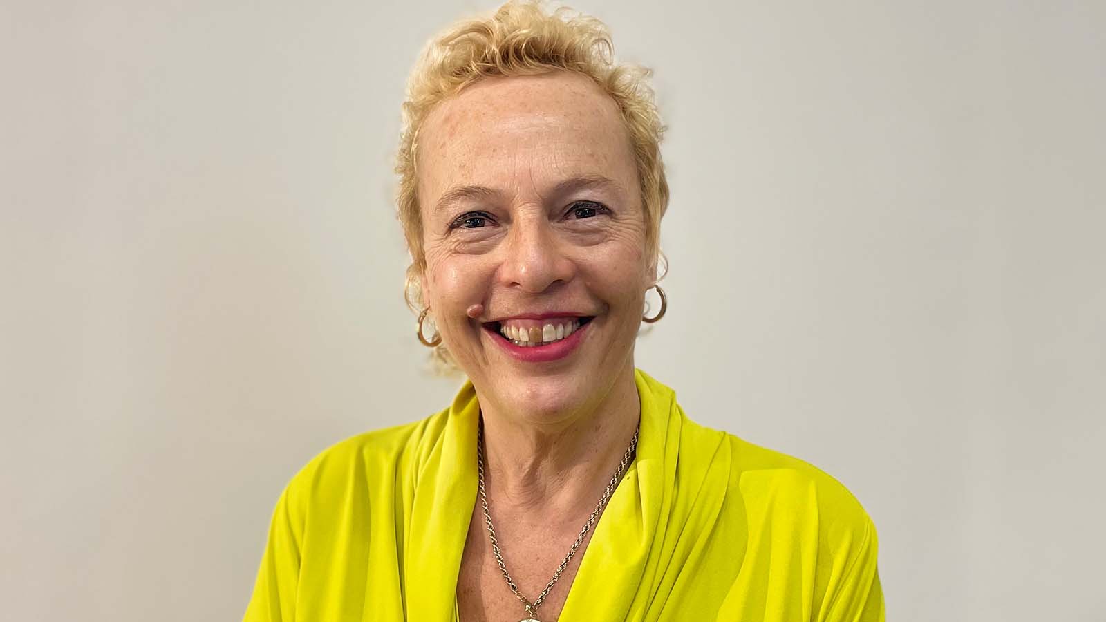 A woman with curly blonde hair wears a bright yellow outfit and a silver necklace, standing against a neutral background. This is Adele Patrick.