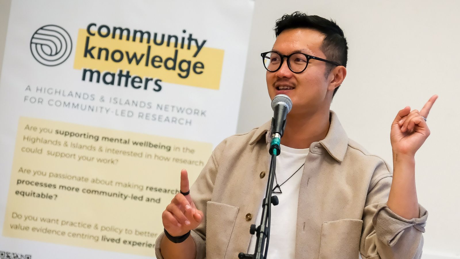 A speaker gestures passionately while presenting next to a banner promoting community-led research and mental wellbeing. This is Lewis Hou.