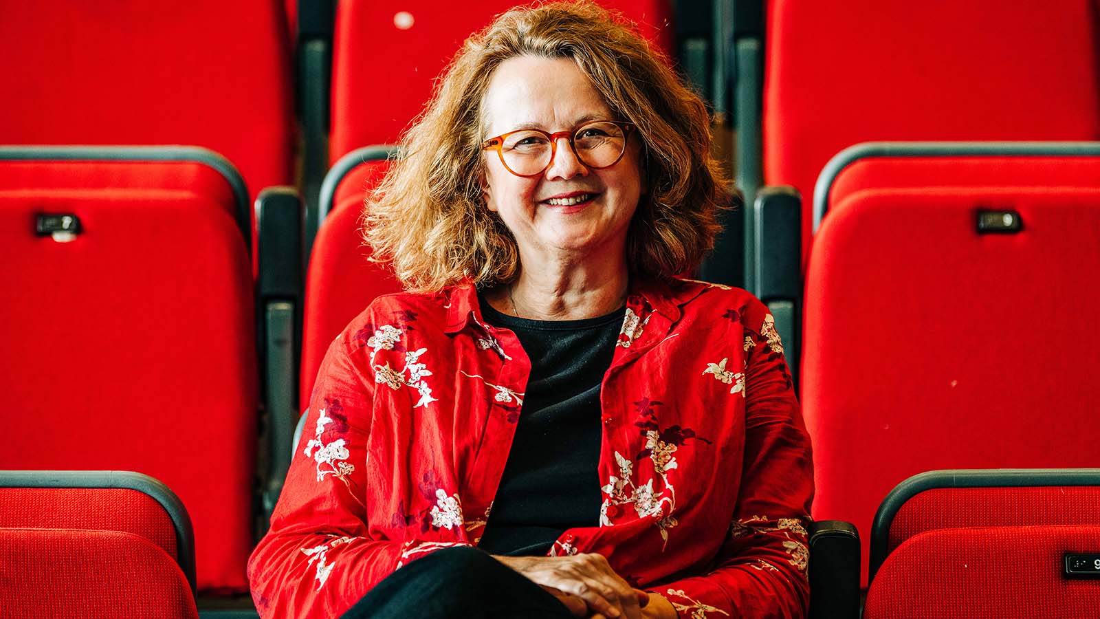 A white woman with short light brown hair, in a bright red patterned shirt sitting in a bank of red theatre seats. This is Magdalena Schamberger.