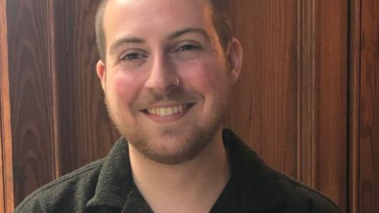A person in a black sweater stands in front of a wooden panel wall, looking at the camera with a neutral expression. This is Milo Clenshaw.