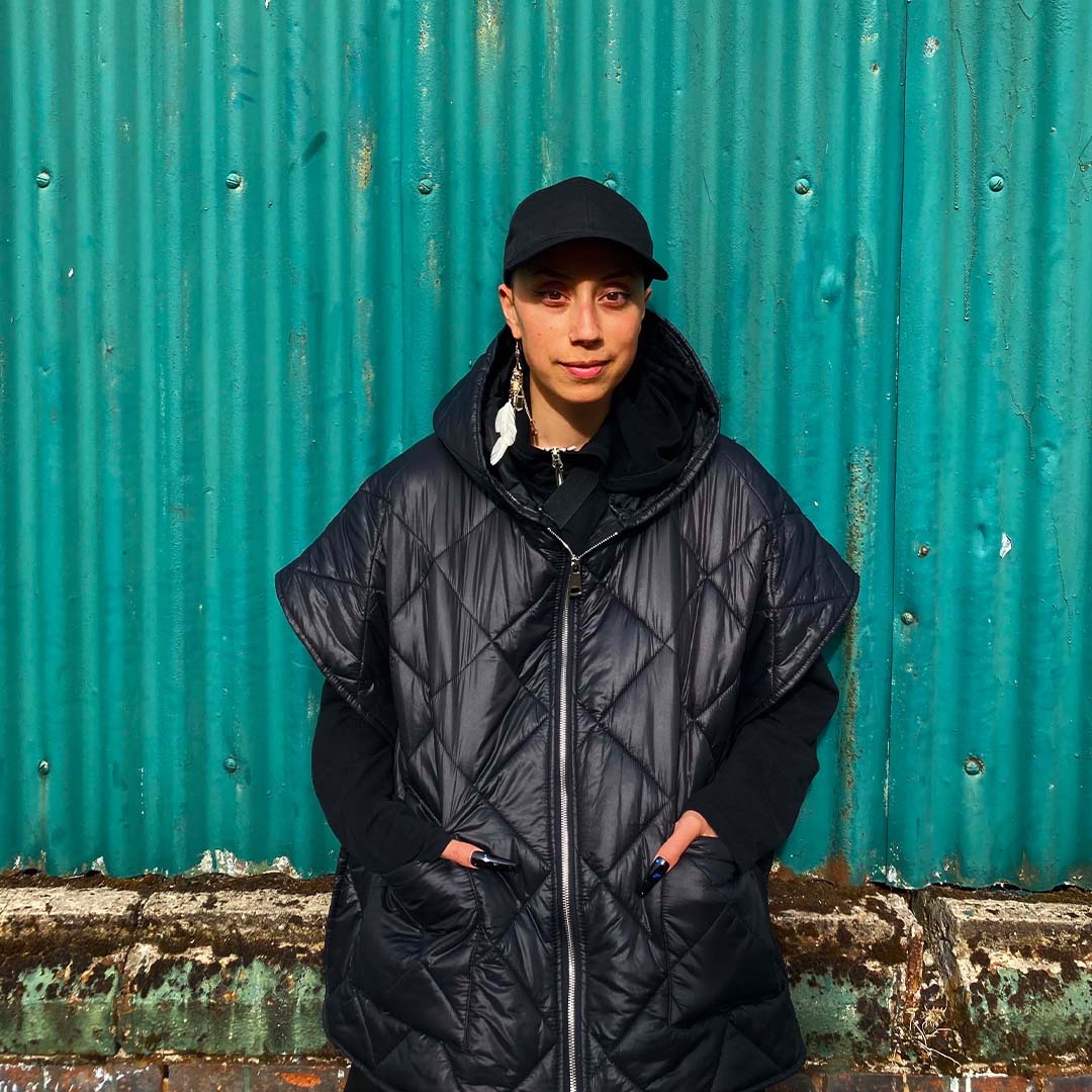 A person wearing an oversized black waterproof coat and black baseball cap, surrounded by a colourful teal corrugated iron and a low stone wall. This is nusa Parinussa.
