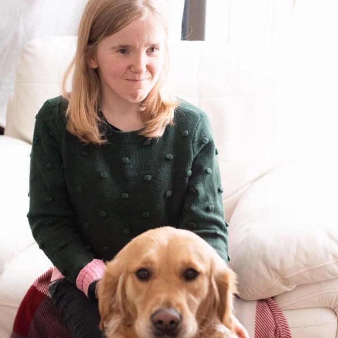 A white woman with short blonde hair wearing a green jumper, next to a Golden Retriever dog. This is Sorcha Pringle.