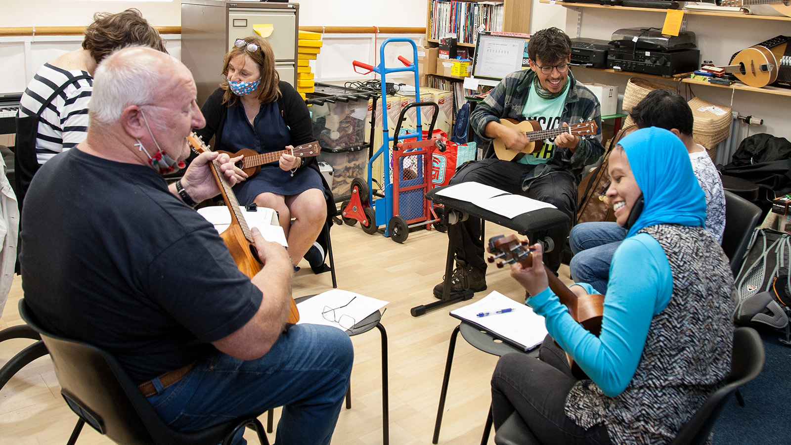 A group of adults sitting in chairs in a circle, facing each other as they each play a ukulele with smiles on their faces and sheet music on their laps