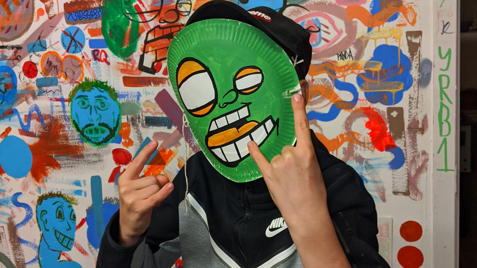 A young man wearing a green mask with a cartoon face on it makes the devil horns hand sign at the camera Behind him is a white wall covered in colourful and playful drawings, illustrations and markings. It’s a visual cacophony