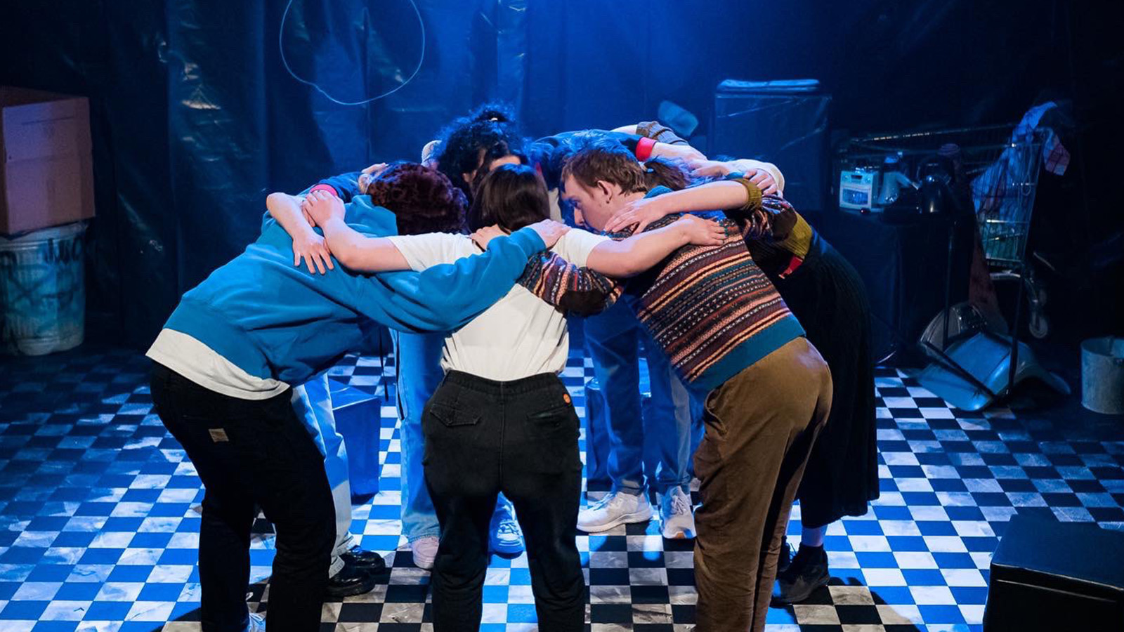 Group of young people stand in a huddle on stage during a performance of Crisis: A Rallying Cry, deep in conversation