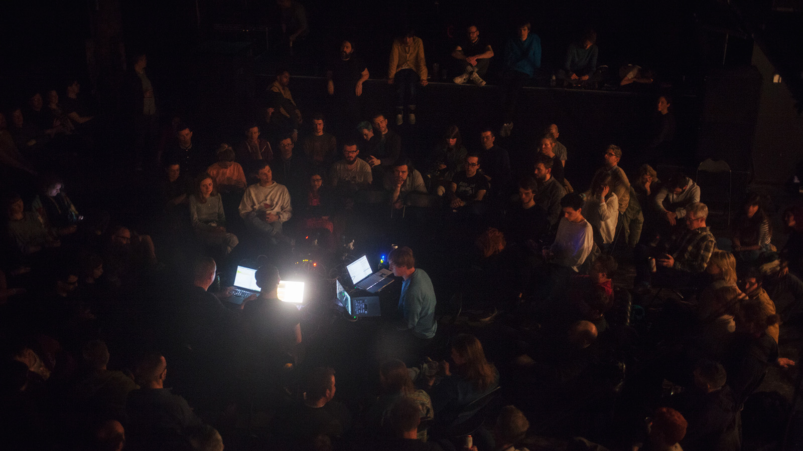A crowd of people gather around a set of four laptops during a set at Counterflows music festival. The light from the devices illuminates the people around them.