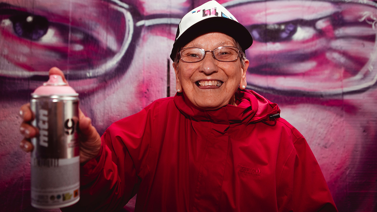 Older woman in bright red jacket smiles as she holds a can of spraypaint aimed at the camera. Behind her is a mural of a woman wearing glasses