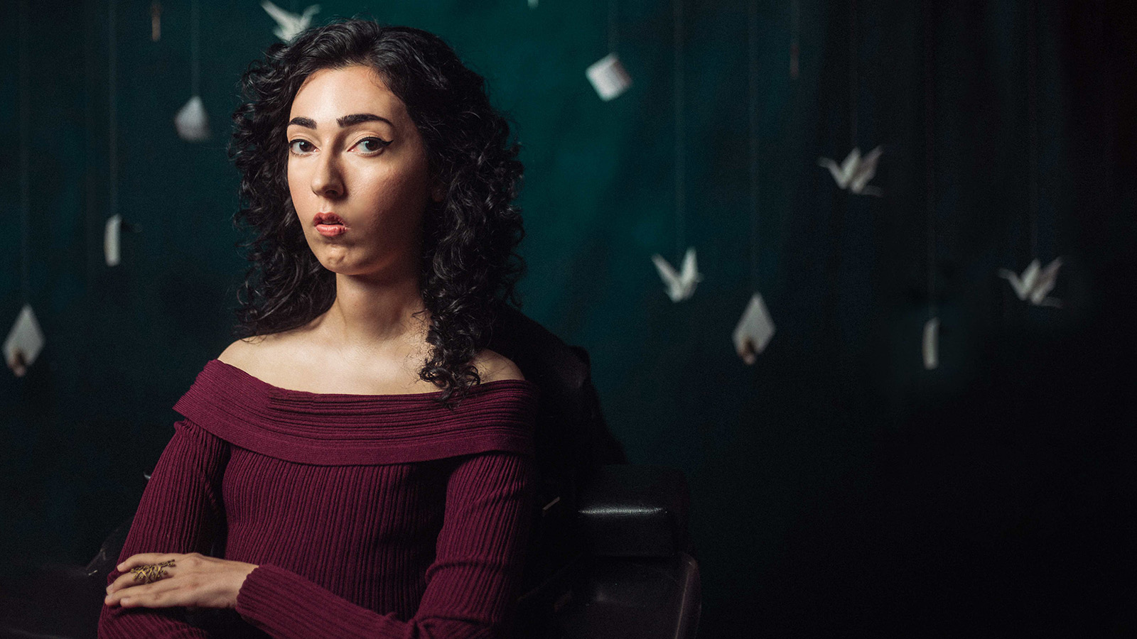 Artist, campaigner and filmmaker Kiana Kalantar-Hormozi sits against a rich blue-green-black background. She faces the viewer with a fixed stare. She is surrounded by envelopes suspended by barely visible string, so they almost look like birds. They appear to be slowly burning, the flames dancing about their edges.