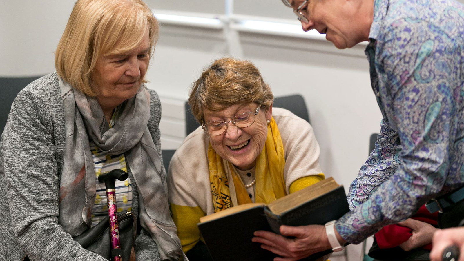 Three older women sit together laughing and reading a book