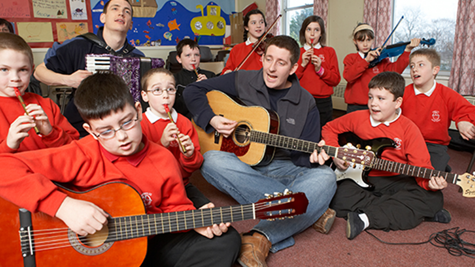 A group of primary-aged schoolchildren in bright red jumpers sit in a classroom playing guitars with two musicians playing the guitar and the accordian
