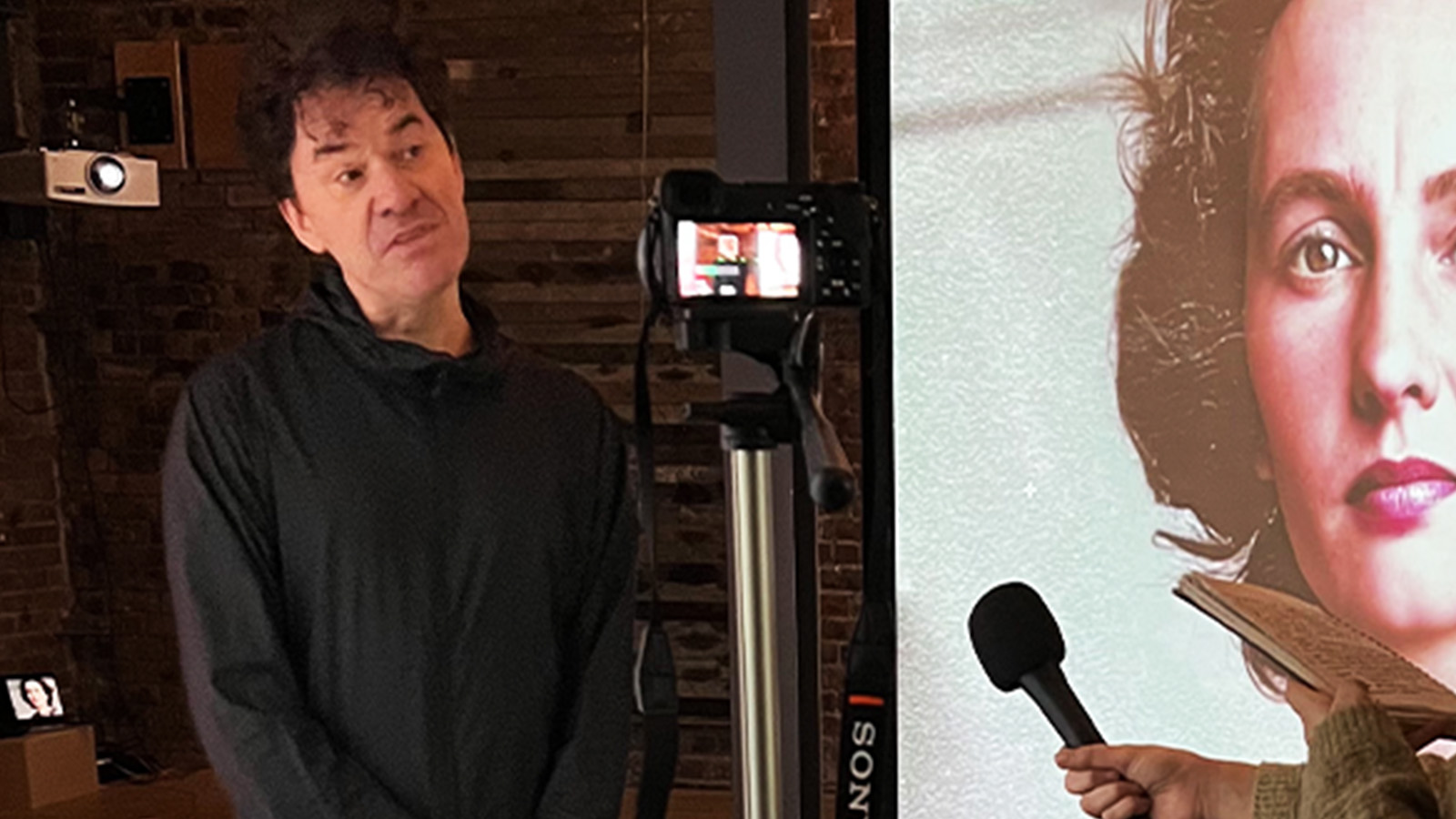 A man with curly black hair stands speaking into a microphone while behind him a huge screen displays an old photograph of the pensive face of a woman