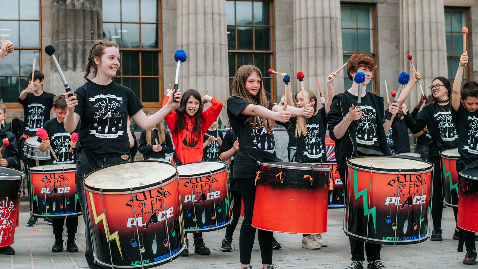 A large group of young people form a band of drummers as they play for audiences outside in Edinburgh