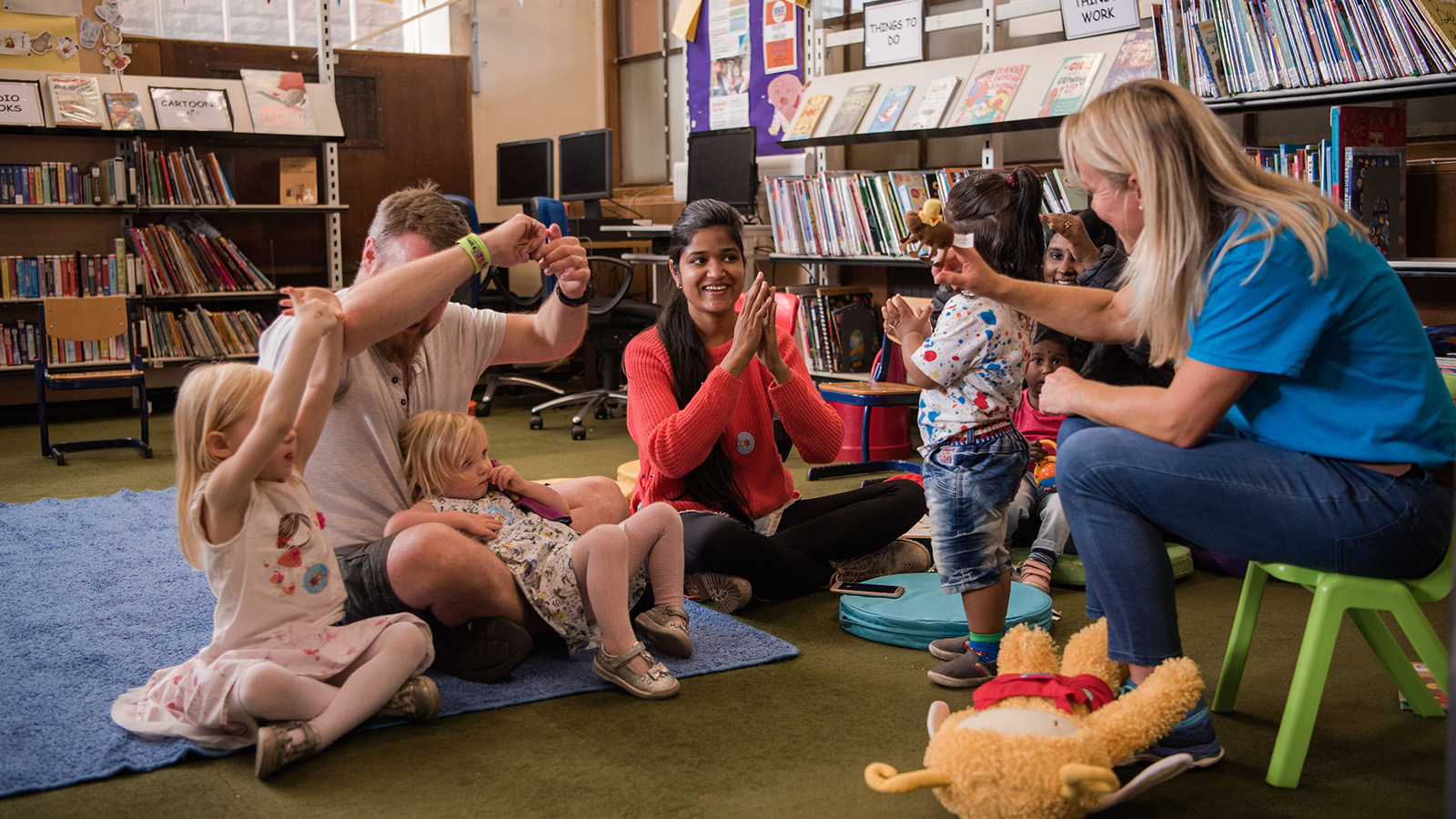 A group of adults and children sit in a circle smiling, laughing and gesturing as they sing a song together in a library.