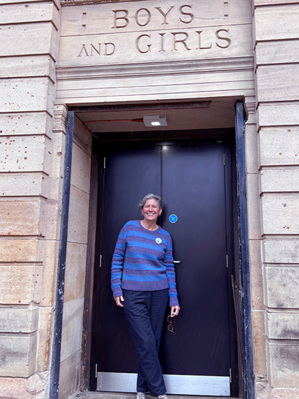 Sue John stands in front of a door of the Glasgow Women's Library which is a Victorian building, the words boys and girls are etched in the stone above her - she is smiling broadly