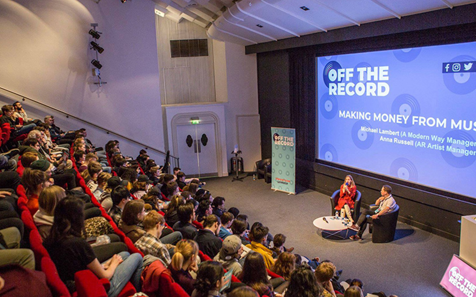 An audience watch a panel of two speakers seated on a stage. A large screen behind them reads Off The Record